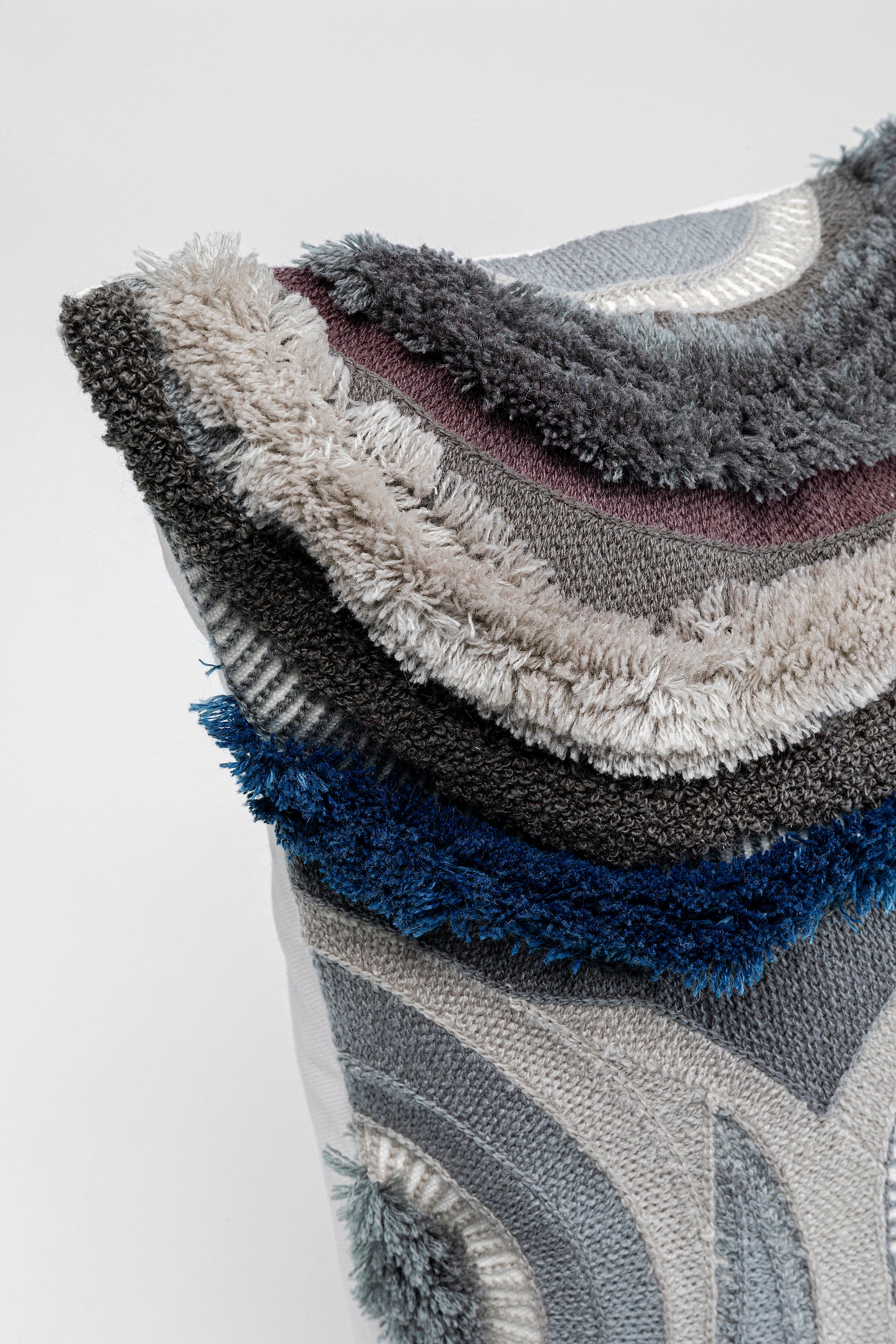 Nahaufnahme eines Kissens mit strukturiertem, mehrfarbigem, geschichtetem Stoff in Grau-, Blau-, Beige- und Schwarztönen.