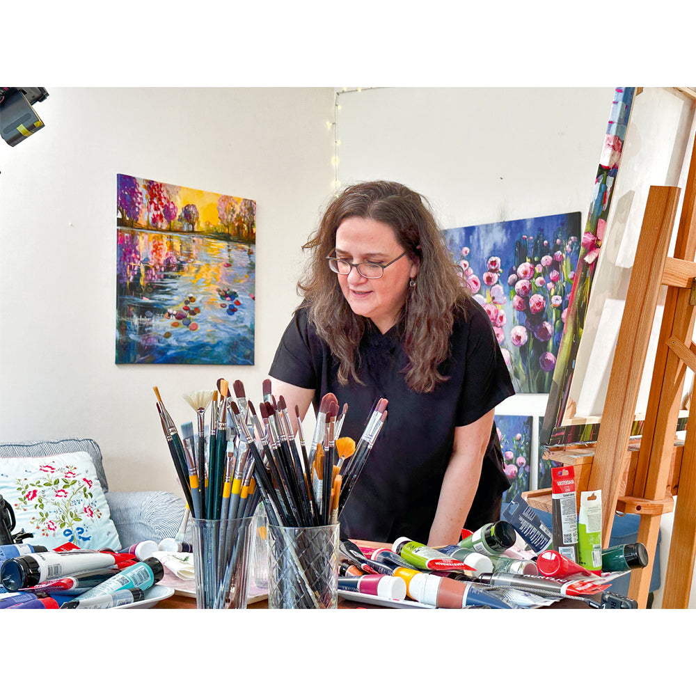 Eine Frau steht mit Pinseln und Farben an einem Tisch, umgeben von farbenfrohen Blumengemälden, darunter Sandra Gebhardt-Höpfners gerahmtes Kunstwerk „My Poppies 1“ (2024, serielles Unikat).