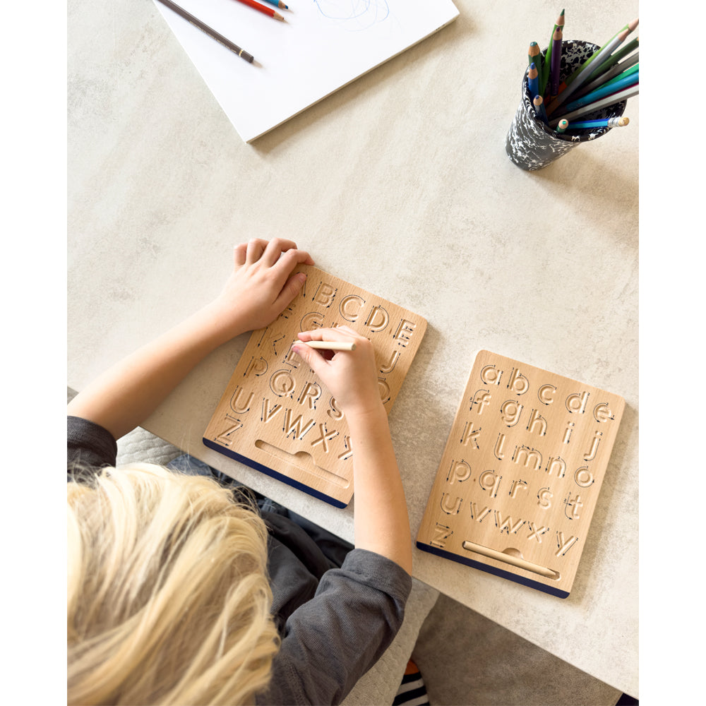 Ein Kind zeichnet Buchstaben des Alphabets auf die Holztafel des Schreibtafel-Sets „Educate“, während Buntstifte und Papier daneben auf dem Tisch liegen.