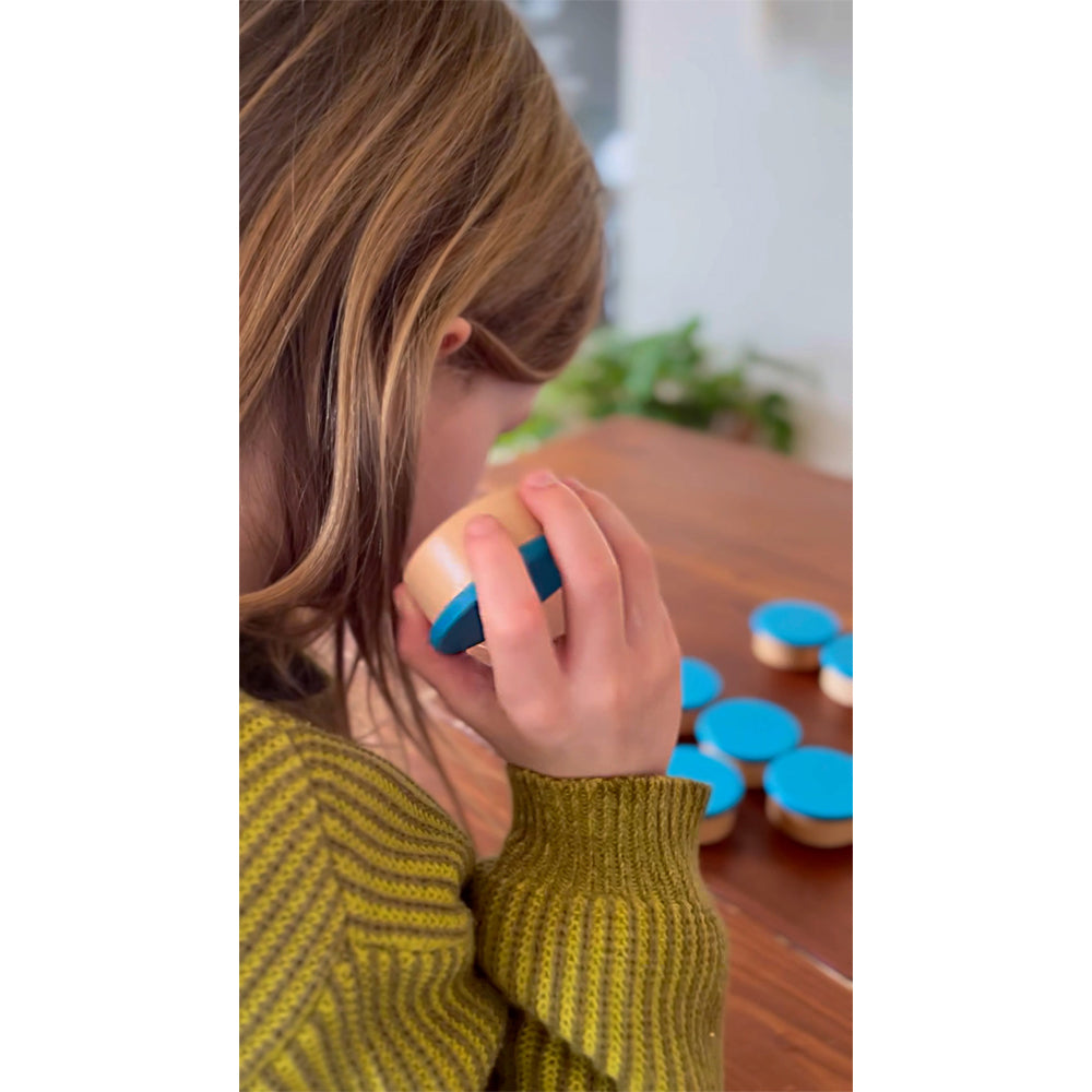 Ein Kind im gelben Pullover hält an einem Tisch mit weiteren ähnlichen Behältern einen Hörmemo-Behälter „Sensory“ mit blauem Deckel ans Ohr.
