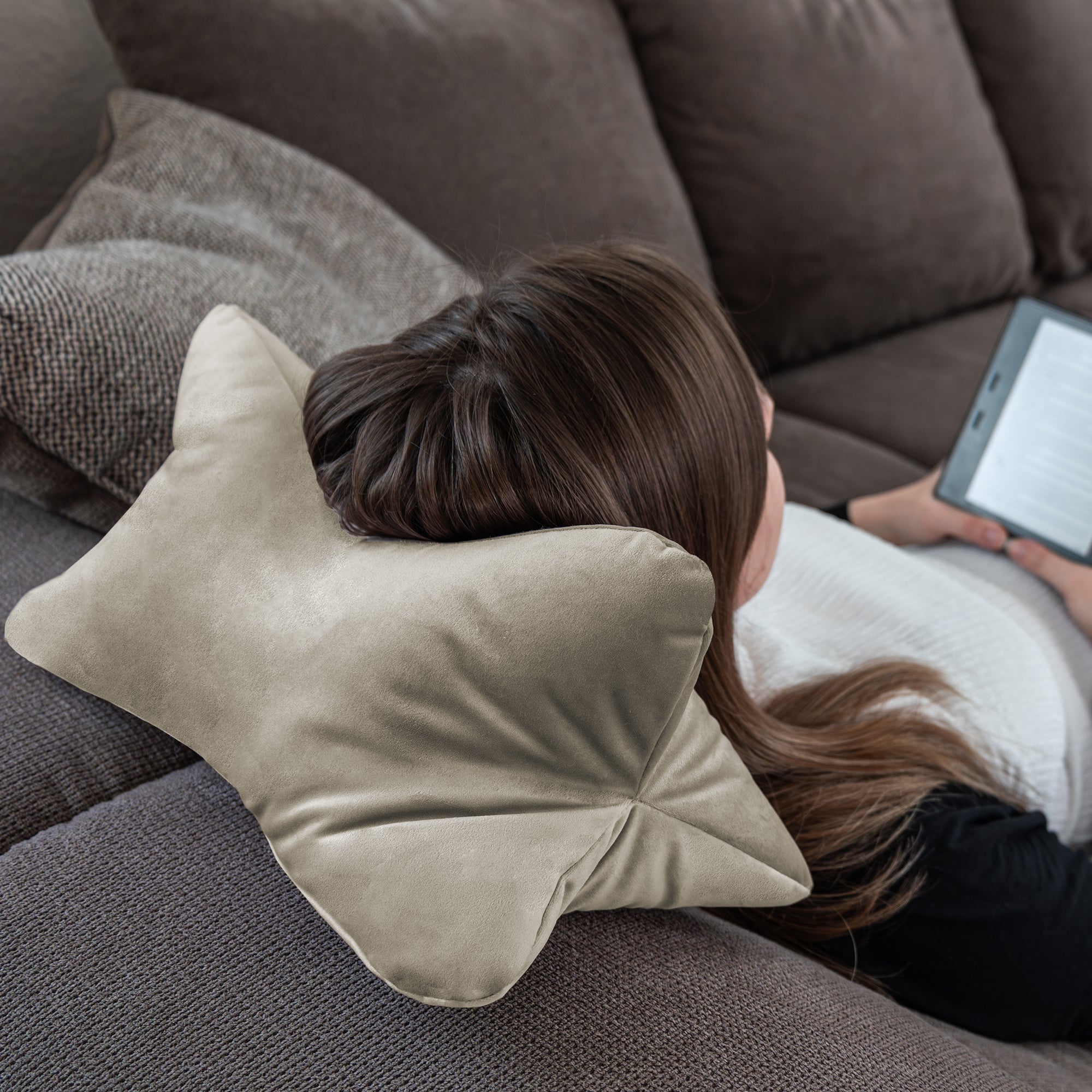 Eine Person lehnt sich mit dem Leseknochen Cozy Samt – Beige, einem schmetterlingsförmigen Kissen mit ergonomischer Nackenstütze, auf einem Sofa zurück und hält einen E-Reader in der Hand.