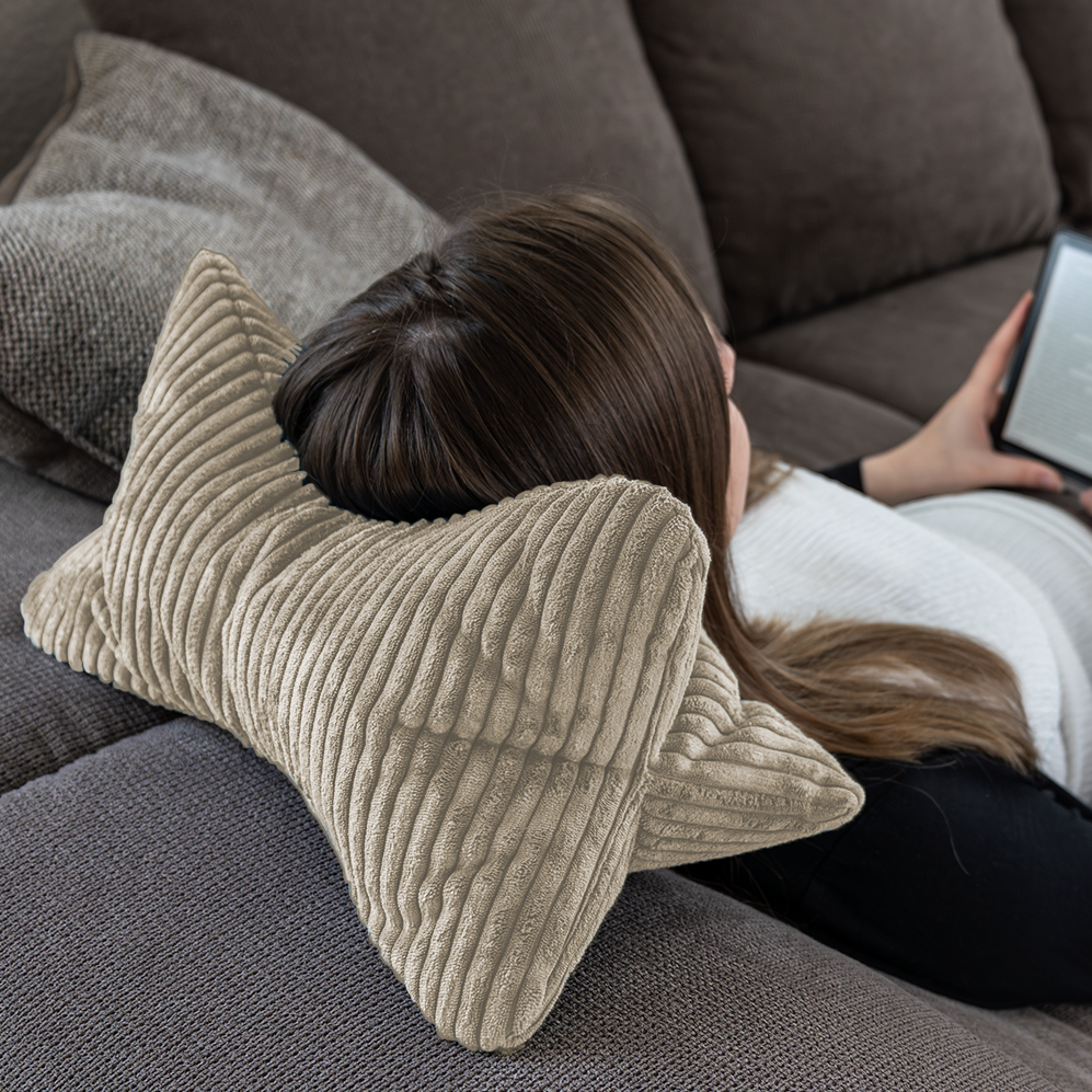 Eine Person lehnt sich auf einem Sofa zurück, legt ihren Kopf auf den beigen Leseknochen Cozy Shara und hält für eine gesunde Körperhaltung einen E-Reader oder ein Tablet in der Hand.