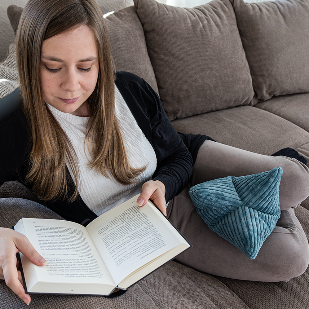 Eine Frau sitzt auf einem grauen Sofa, liest ein Buch mit dem Leseknochen Cozy Shara – Petrol als Nackenstütze und genießt optimalen Komfort und Entspannung.
