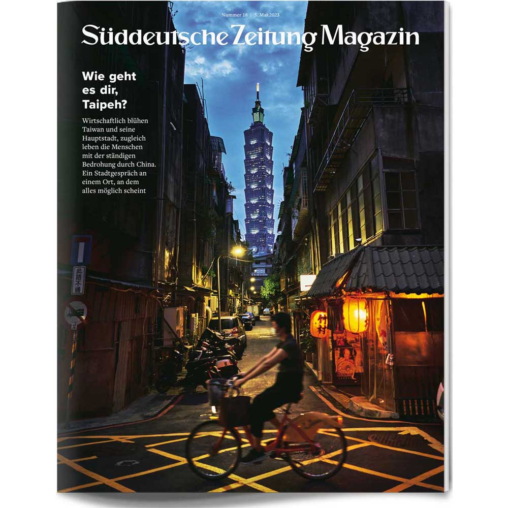Eine Person fährt in der Abenddämmerung mit dem Fahrrad eine enge Straße in Taipeh entlang, während im Hintergrund der Turm Taipei 101 beleuchtet ist.