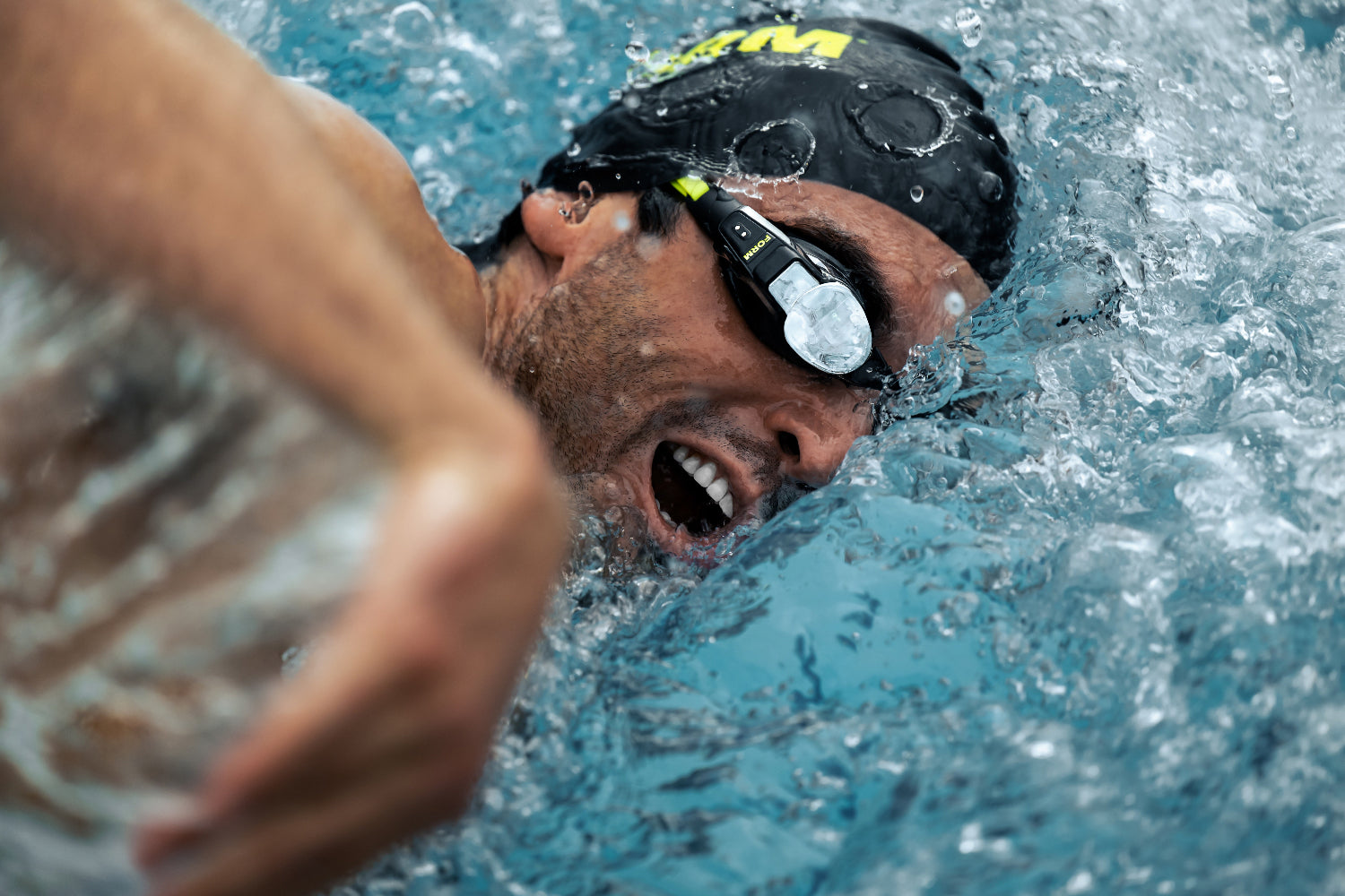 Ein Schwimmer mit schwarzer Kappe und Schwimmbrille führt einen Freistilschwimmzug in einem Schwimmbecken aus, wobei das Wasser um sein Gesicht spritzt.