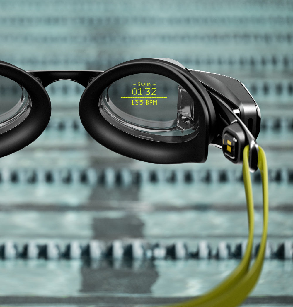Nahaufnahme einer intelligenten Schwimmbrille, die Schwimmzeit und Herzfrequenz auf einem digitalen Bildschirm anzeigt, mit einem Schwimmbad im unscharfen Hintergrund.