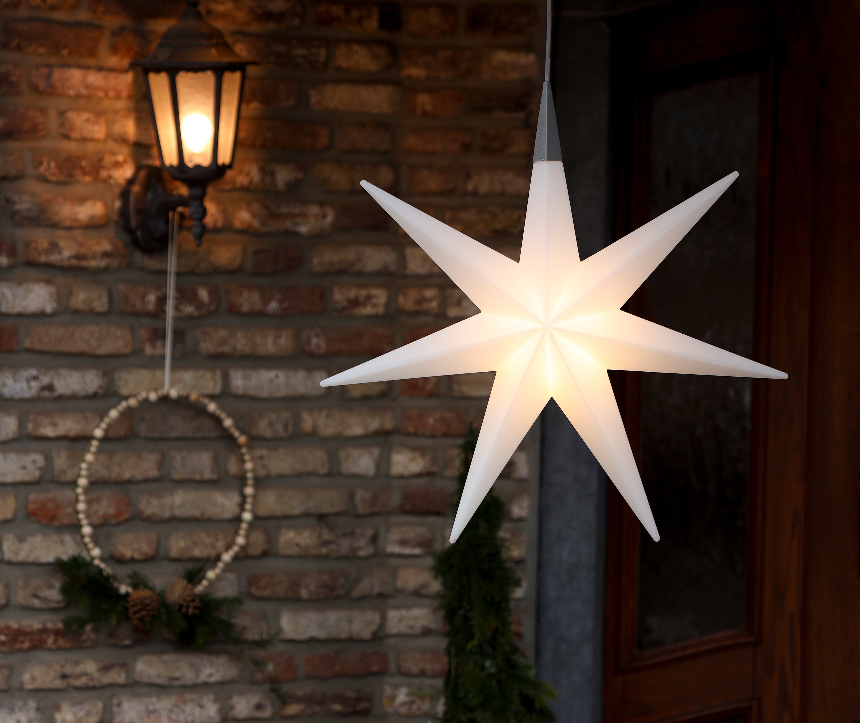 Leuchtende, sternförmige Laterne, die an einer Ziegelwand hängt, neben einer brennenden Lampe und einem einfachen Kranz.