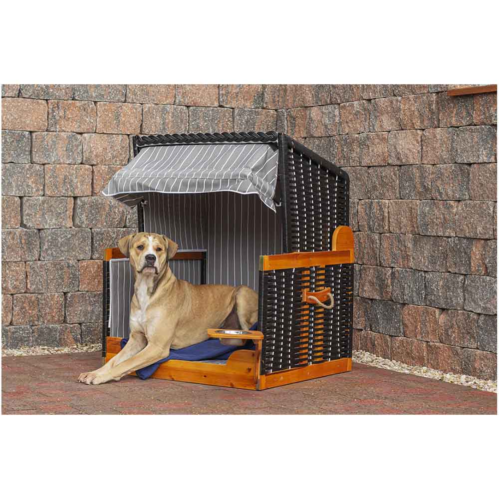 Ein großer Hund sitzt auf einem Kissen in einer Hundehütte aus Weidengeflecht mit eingebautem Vordach vor einem Hintergrund aus Steinmauern.