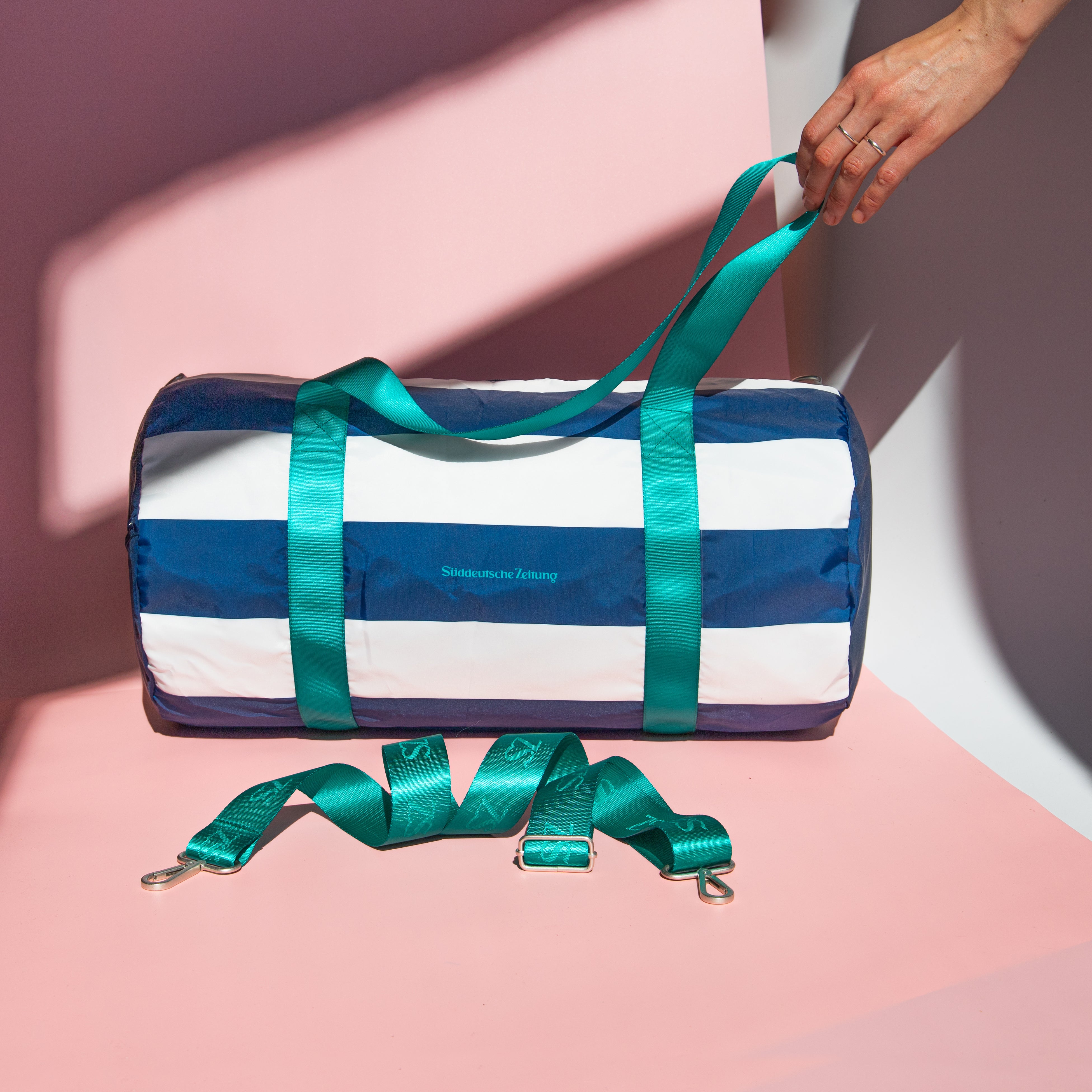 Tasche Weekender blau weiß gesreift mit grünen Henkeln und Logo Süddeutsche Zeitung auf rosa Hintergrund. Eine Hand im Bild hält die Henkel der Tasche