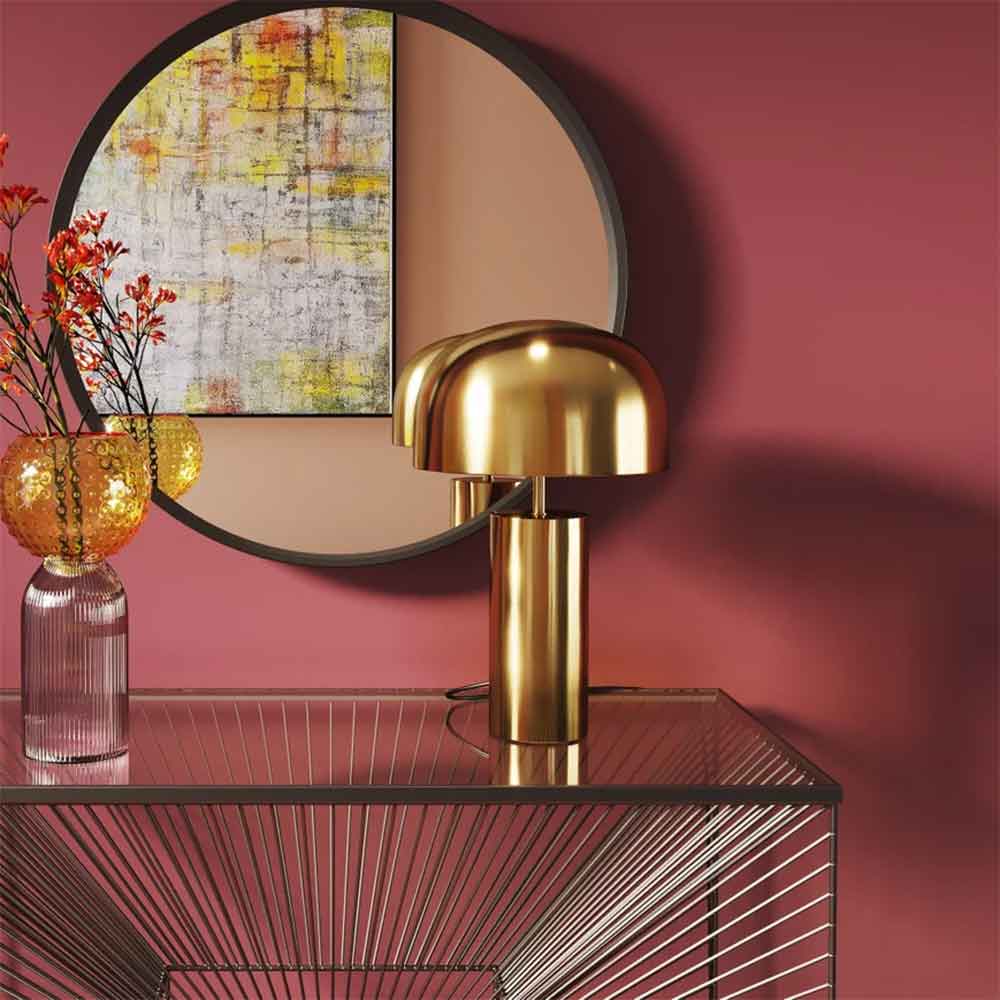Eine goldene Lampe auf einem Glastisch, ein runder Spiegel, abstrakte Kunst und eine Blumenvase vor einer rosa Wand.