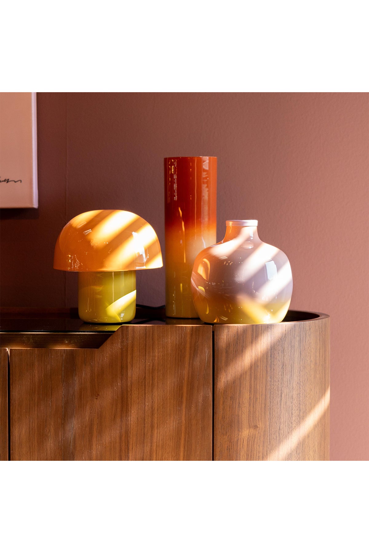 Drei dekorative Glasobjekte, darunter eine pilzförmige Lampe und zwei Vasen in Gelb- und Orangetönen, sind auf einer Holzvitrine vor einer malvenfarbenen Wand angeordnet, auf die das Sonnenlicht Schatten wirft.