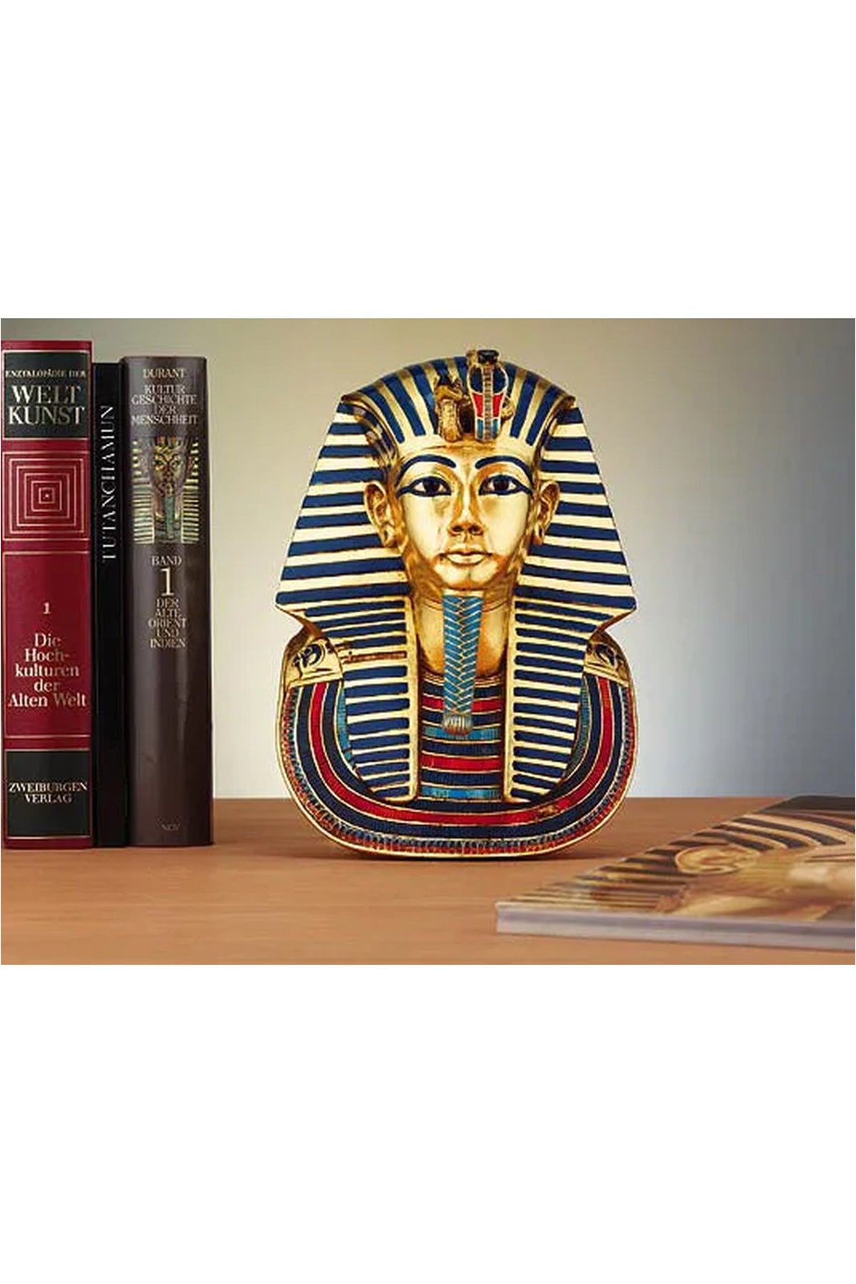 Ein Modell der goldenen Maske von König Tutanchamun steht auf einem Holzregal zwischen Büchern, darunter auch Bände zur Kunstgeschichte, wobei ein weiteres Buch teilweise im Vordergrund zu sehen ist.