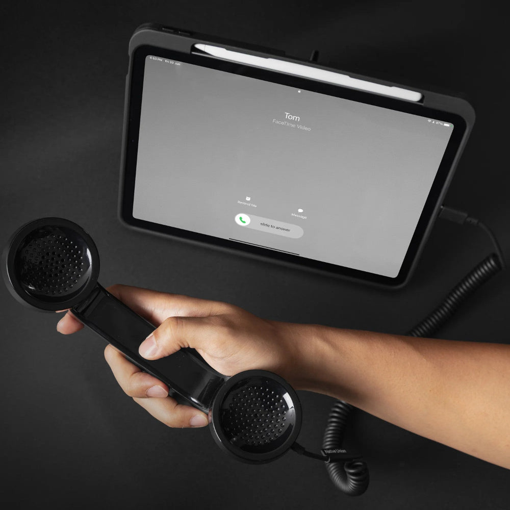 Eine Hand hält ein Retro-Telefon, das mit einem Tablet verbunden ist und einen eingehenden FaceTime-Videoanruf von Tom vor einem schwarzen Hintergrund anzeigt.