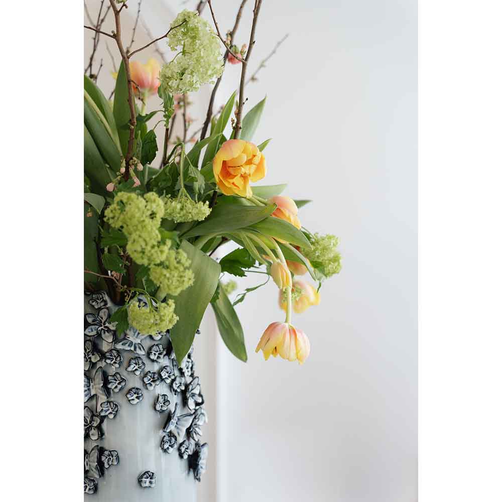 Gelbe Tulpen und grüne Blumen in einer strukturierten weißen Vase vor einem schlichten, hellen Hintergrund.