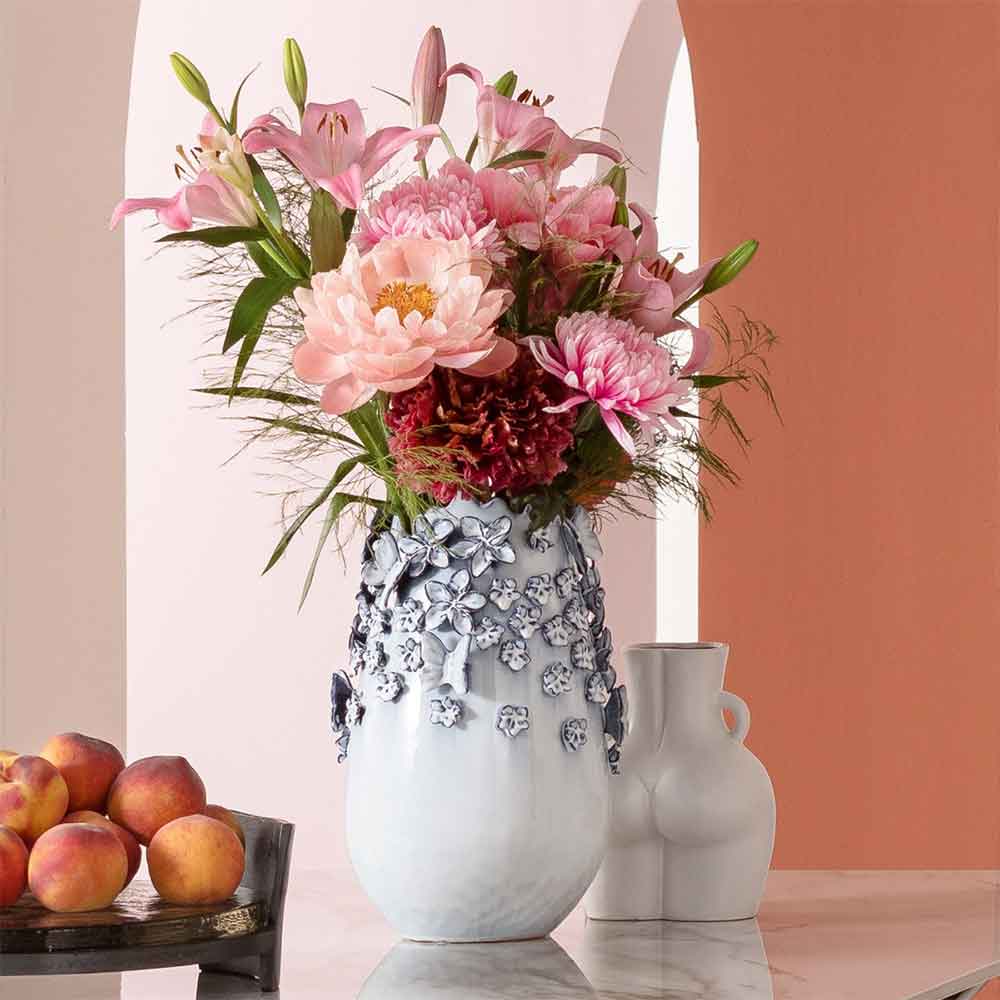 Eine Vase mit rosa Blumen auf einem Marmortisch, daneben Pfirsiche und ein weißer Zierkrug.