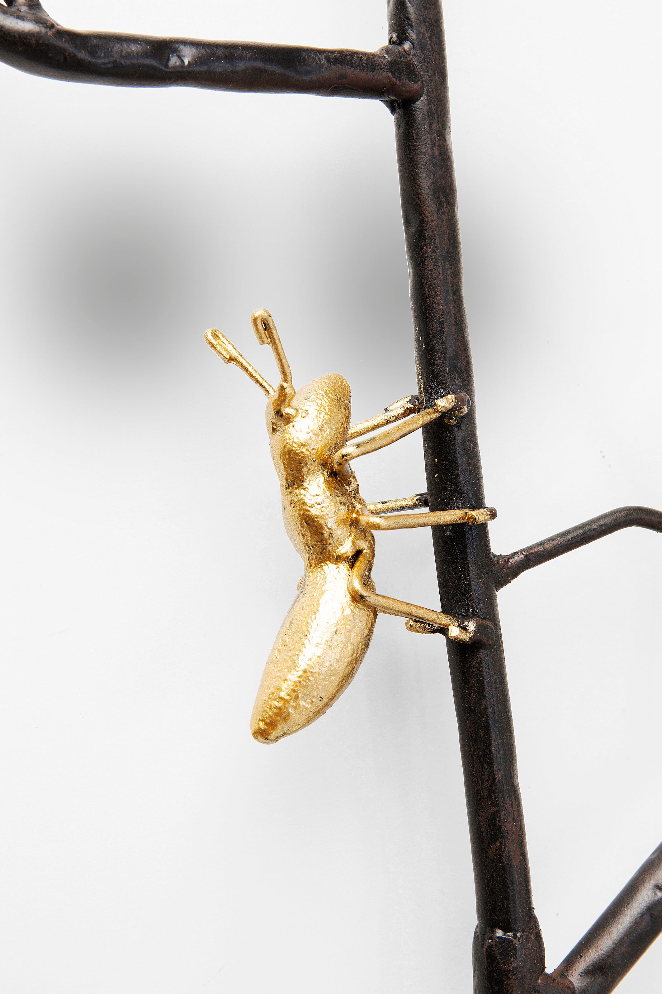 Eine goldfarbene Ameisenskulptur, die auf einem dunklen Metallzweig vor einem weißen Hintergrund klettert.