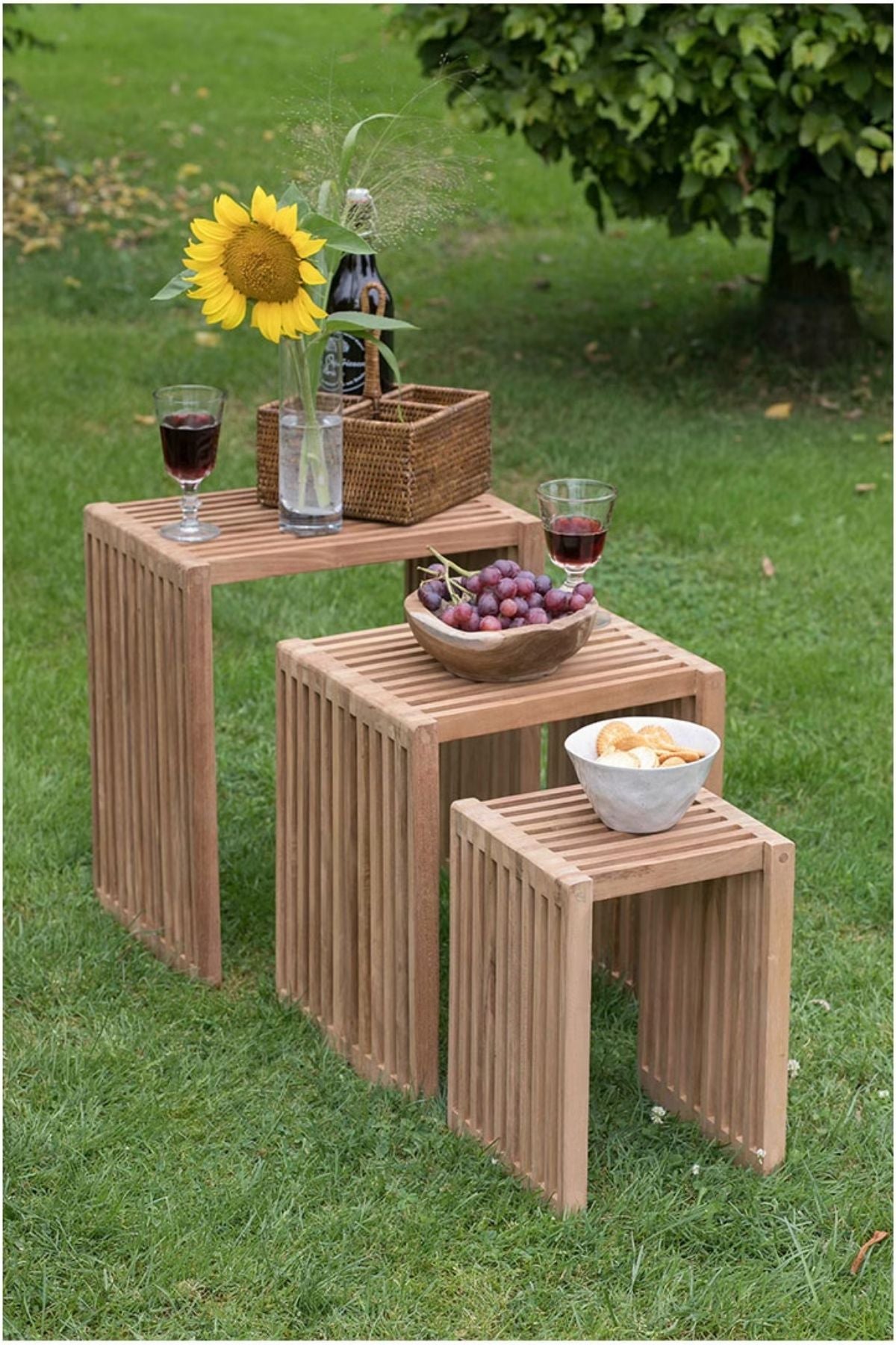 Auf drei ineinander gesteckten Holztischen auf dem Rasen stehen Wein, Gläser, eine Sonnenblume in einer Vase, ein Korb, Weintrauben und eine Schale mit Chips, im Hintergrund ist ein Baum zu sehen.