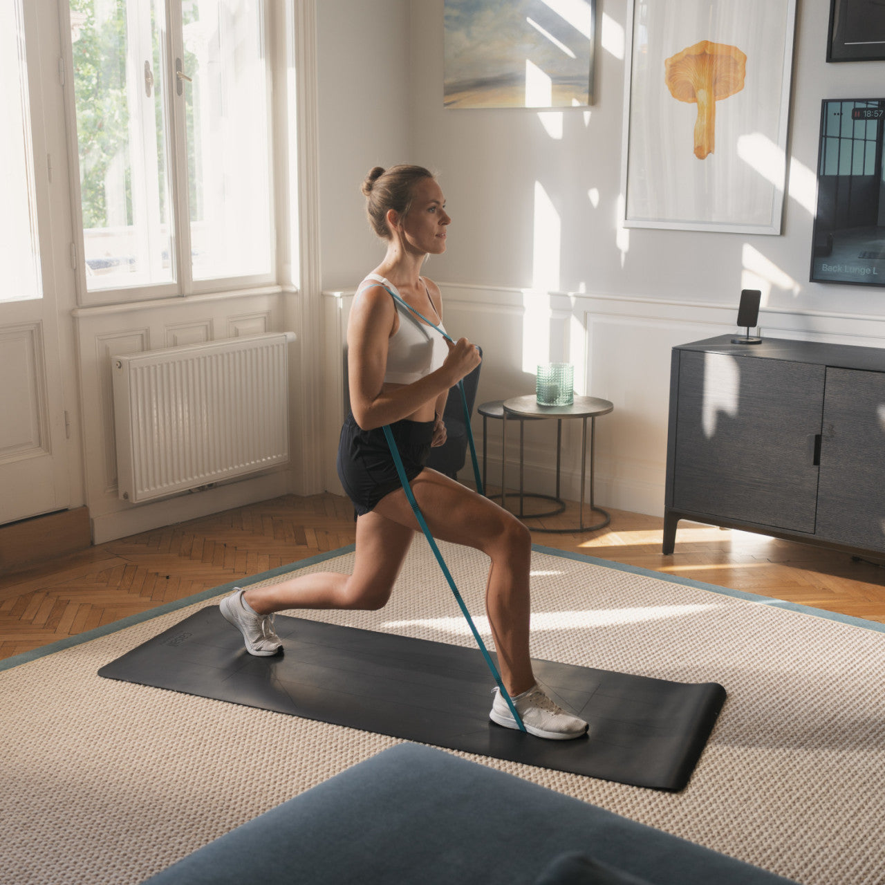 Eine Frau trainiert drinnen auf einer Matte und macht Ausfallschritte mit dem Smartes Fitnessband STRAFFR mit App - mittel in einem hellen Wohnzimmer.