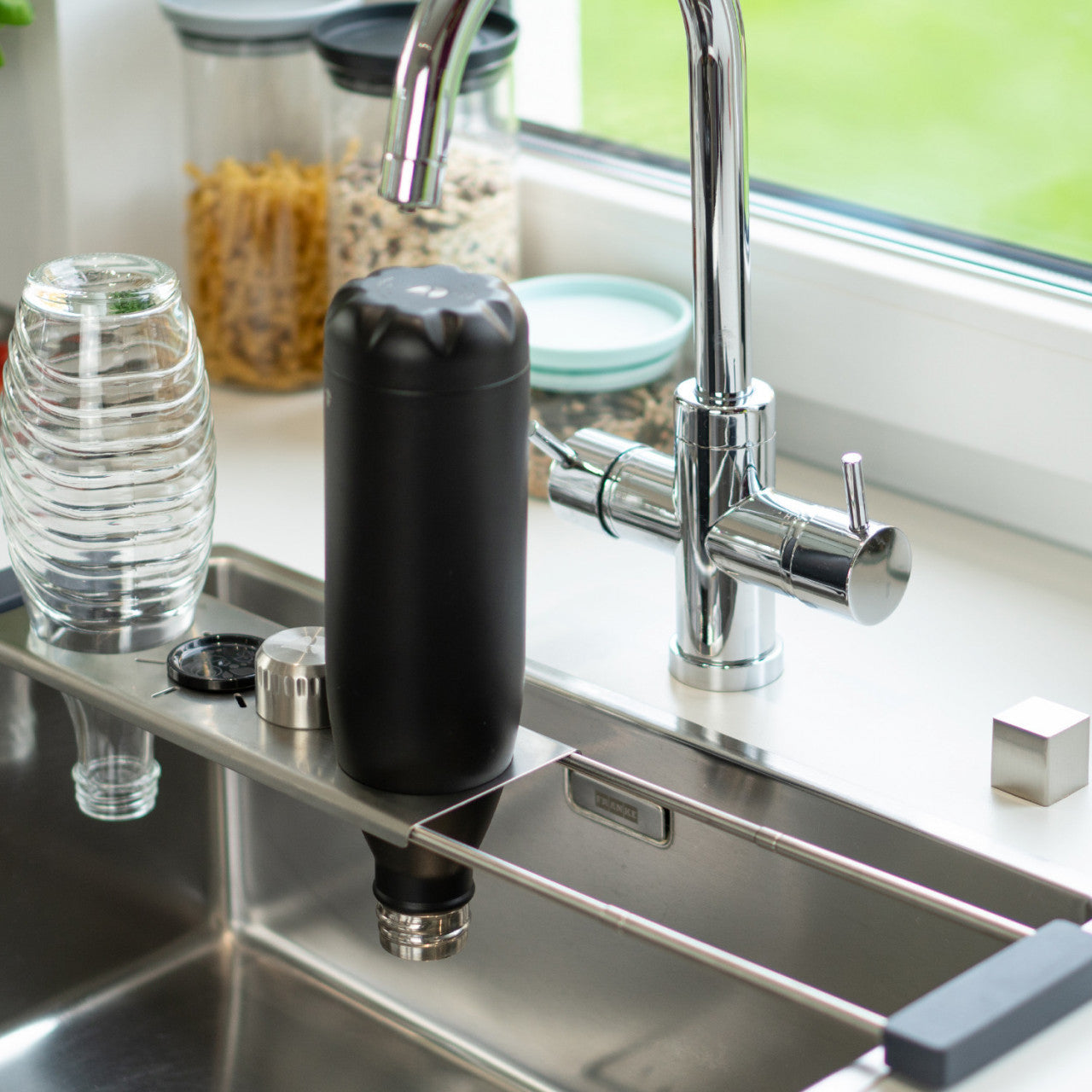 Die schwarze, wiederverwendbare Abtropfhilfe Drip.it-Flasche trocknet kopfüber auf einem Küchenspülenregal neben einem Glas und Gläsern in der Nähe eines modernen Wasserhahns.