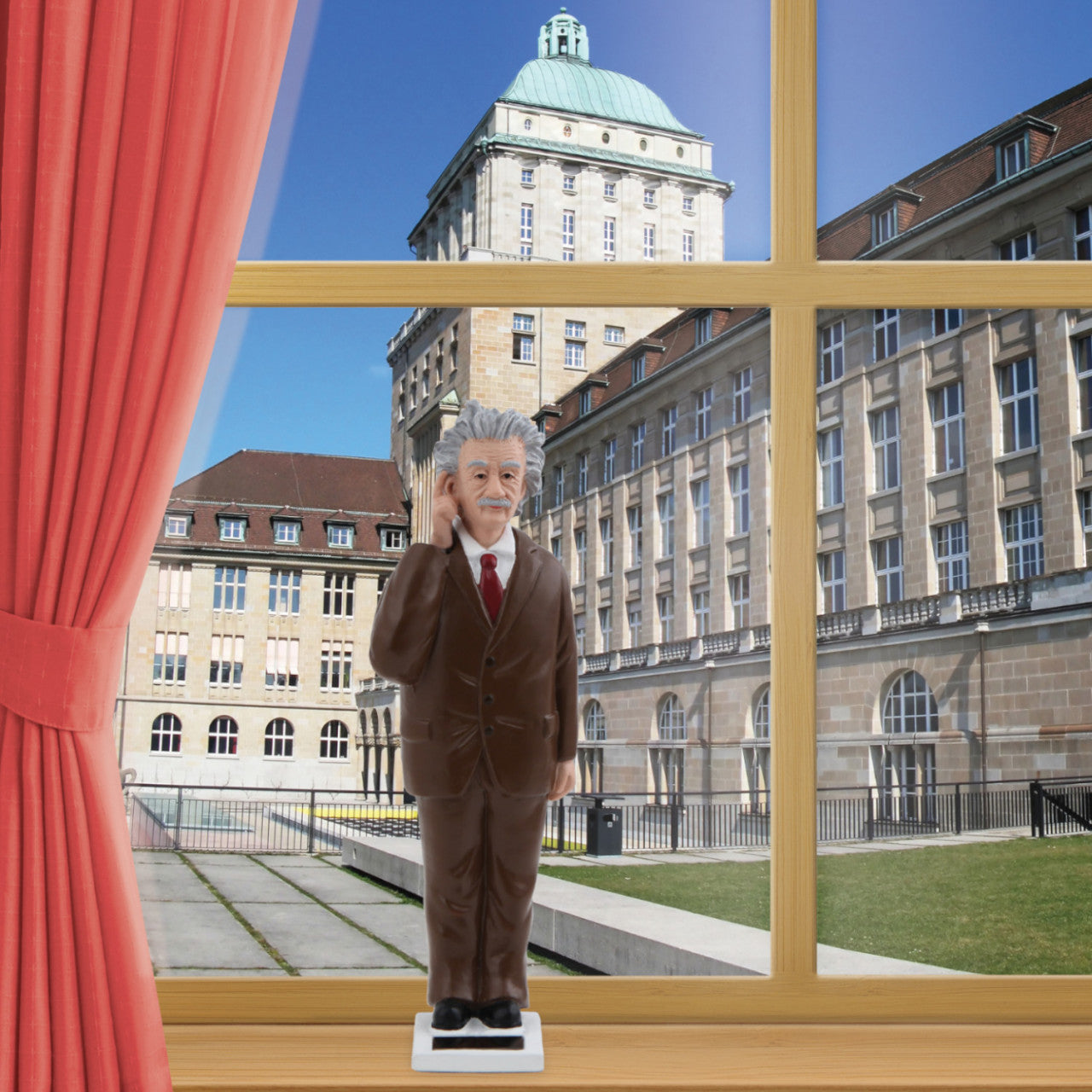 Die Dekofigur Einstein mit Solarantrieb steht an einem Fenster, von dem aus man ein stattliches Gebäude sehen kann.