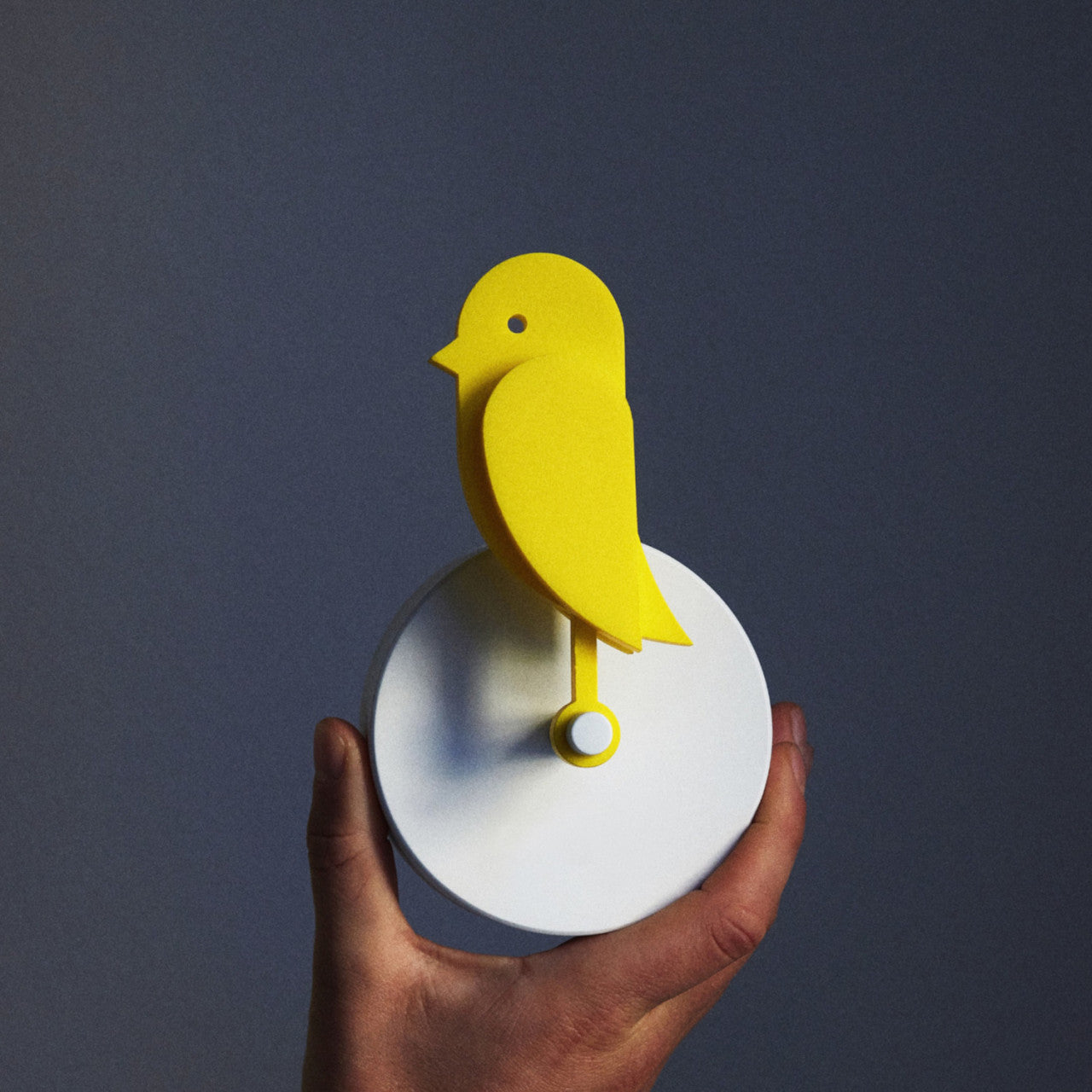 Eine Hand hält den Frischluftmonitor Birdie 2.0 mit CO2-Sensor, ein gelbes, vogelförmiges Gerät auf einem weißen, runden Sockel, vor einem grauen Hintergrund.