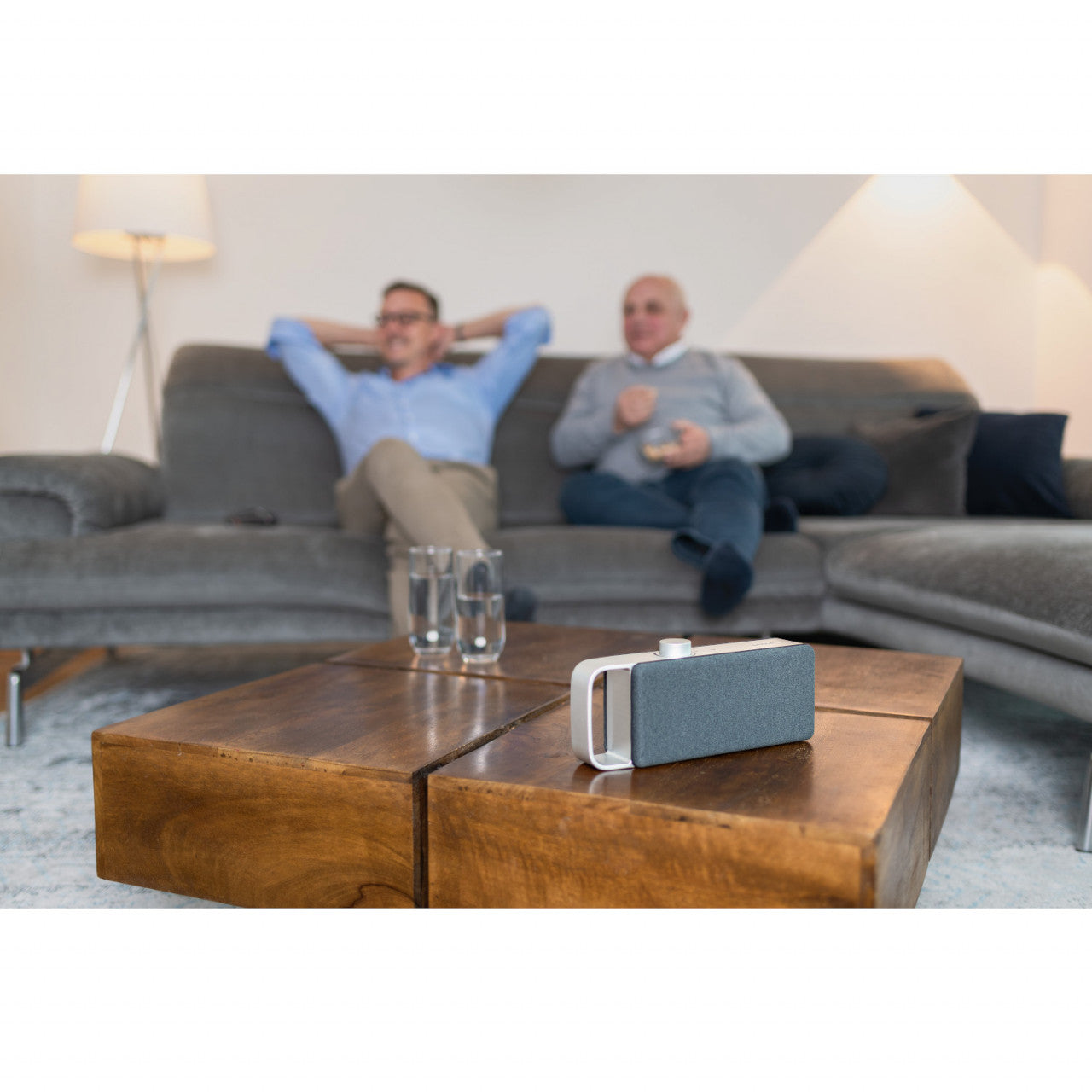 Zwei Männer entspannen auf einem Sofa und sehen fern. Auf dem hölzernen Couchtisch davor stehen der tragbare TV-Sprachverstärker OSKAR und zwei Gläser.