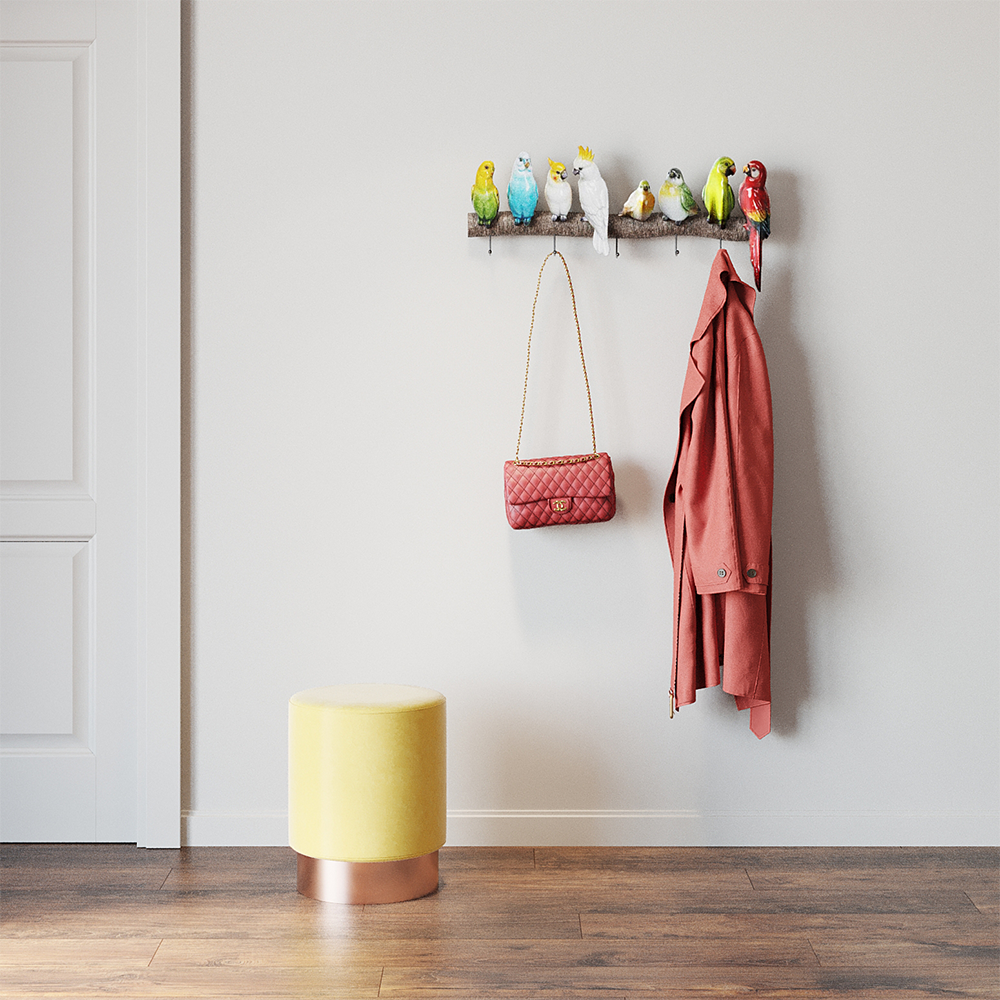 Die Wandgarderobe Exotic Birds mit bunten Kleiderhaken in Vogeloptik enthält eine rote Tasche und einen Korallenmantel. Daneben steht ein gelber Ottoman mit Metallfuß auf Holzboden – pure Sommerstimmung an der hellen Wand.