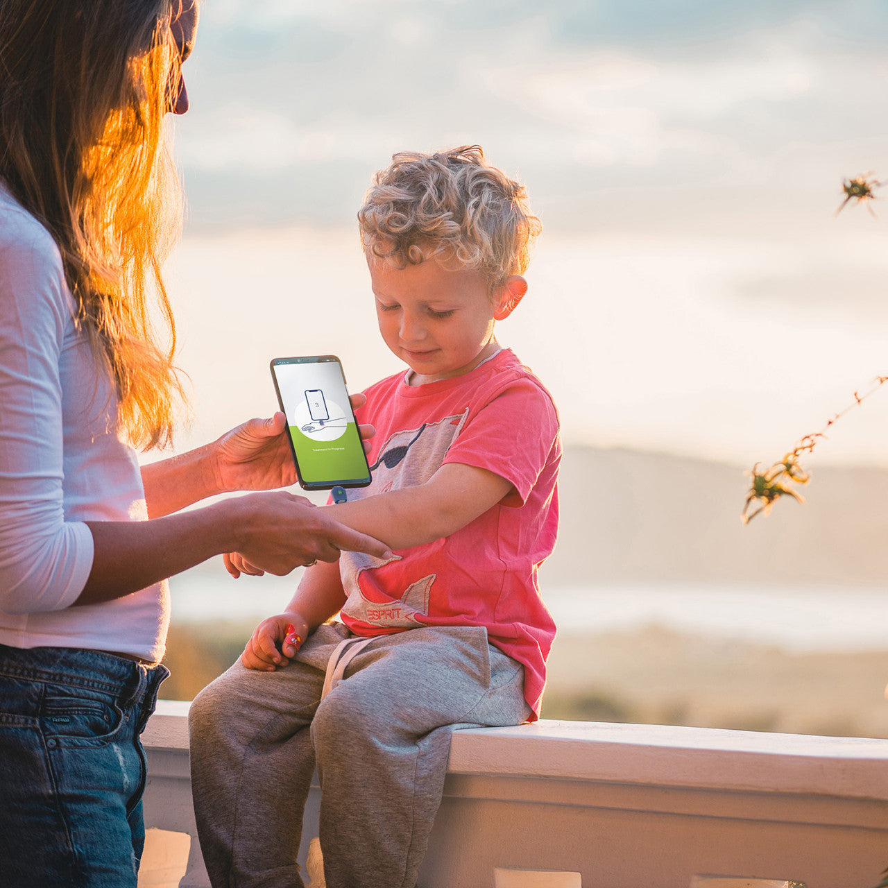 Eine Frau behandelt bei Sonnenuntergang im Freien mit ihrem Smartphone den Arm eines kleinen Jungen mit dem Insektenstichheiler heat it classic Lightning.