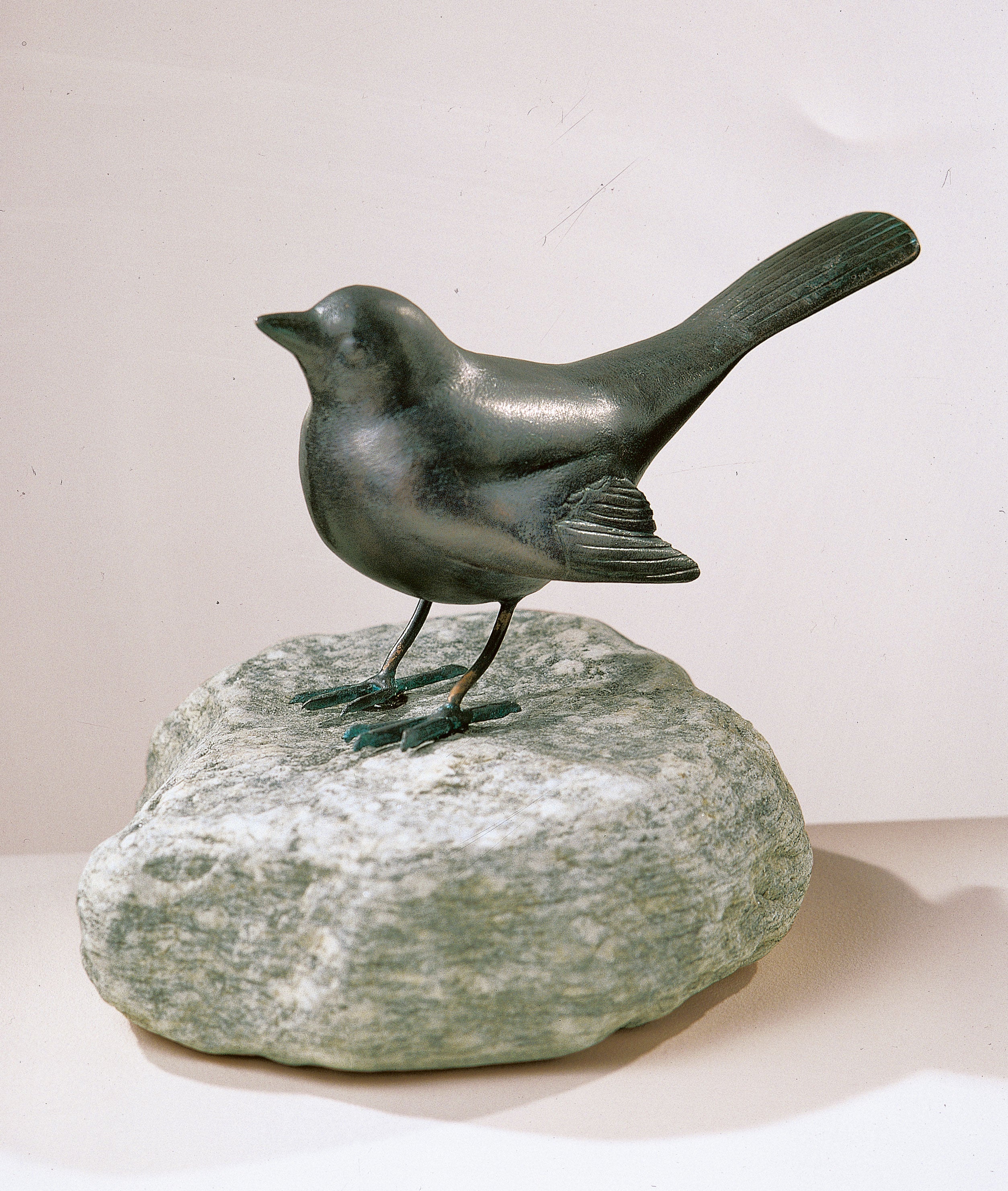 Eine bronzene Vogelskulptur steht auf einem groben Steinsockel vor einem schlichten Hintergrund.