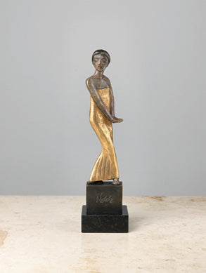 Bronzeskulptur einer stehenden Frau in einem langen Kleid auf einem schwarzen rechteckigen Sockel vor einem schlichten Hintergrund.