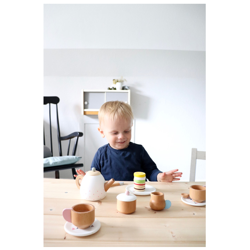 Ein Kleinkind lächelt glücklich, während es an einem Tisch mit dem hölzernen Teeservice und Spielzeug-Macarons von Teeservice Kinderküche spielt.