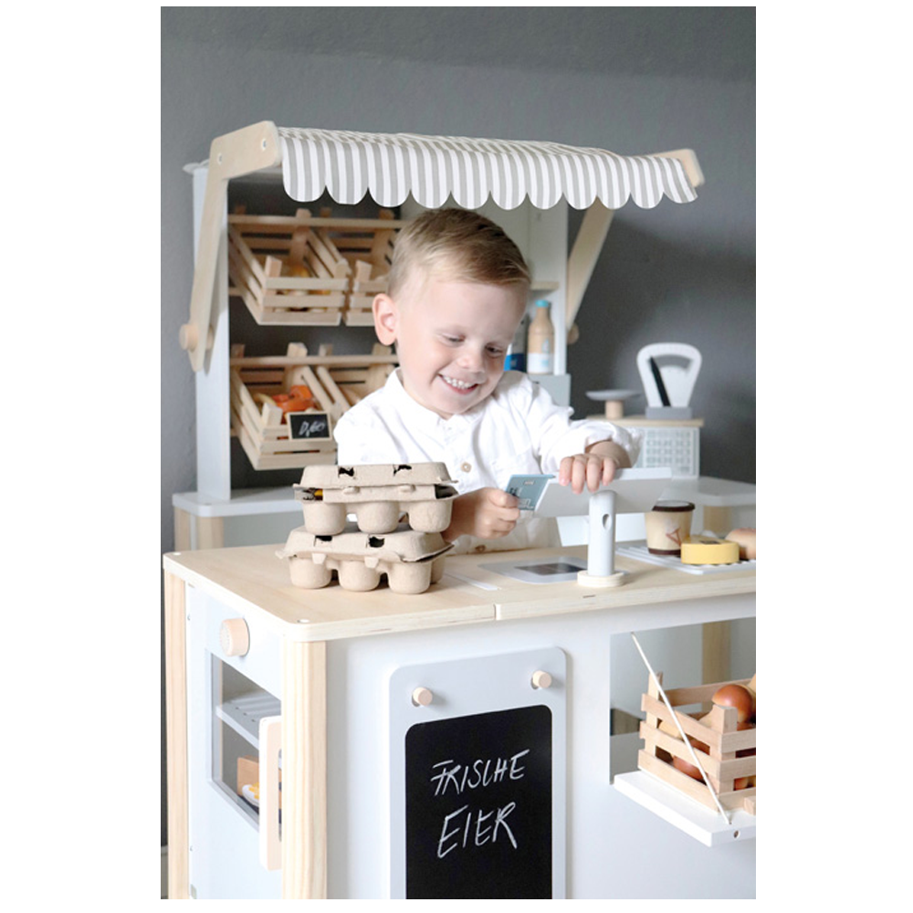 Ein lächelndes Kind spielt mit dem Kaufladen und der Kinderküche fresh, die einen Spielzeug-Marktstand, Eierkartons und eine Spielkasse umfasst.