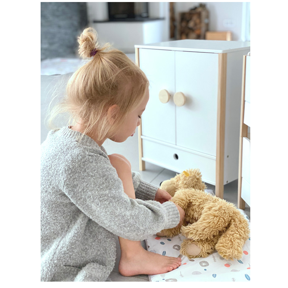 Ein kleines Kind in einem grauen Pullover sitzt auf dem Boden und spielt mit einem Plüschteddybär neben dem weißen Puppenschrank „Little Button“.