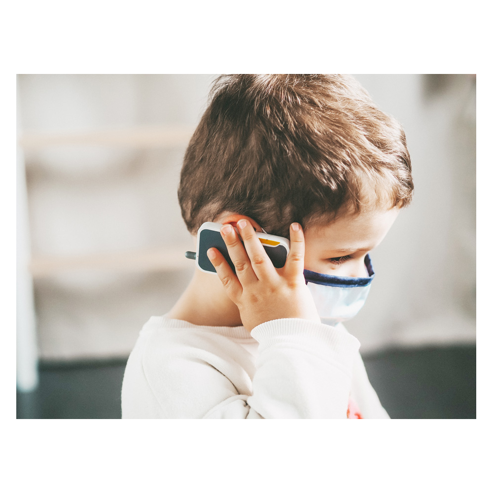 Ein kleines Kind trägt eine Gesichtsmaske und sieht nachdenklich aus, während es einen Notarztrucksack und ein Spielzeugtelefon ans Ohr hält.