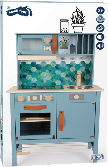 Die Kinderküche „tasty“ ist eine blaue Spielzeugküche aus Holz mit Backofen, Spüle, Herd und einer sechseckigen blaugrünen Fliesenrückwand.