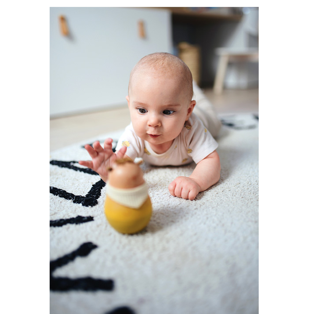 Ein Baby liegt auf dem Bauch auf einem Teppich und greift neugierig nach dem Baby Stehaufspielzeug „Seaside“.