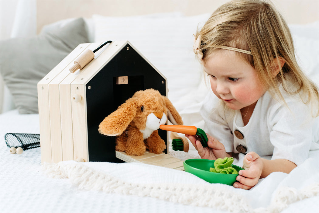 Ein junges Mädchen spielt auf einem Bett mit einem Hasenstall mit Auslauf und füttert ihr Stoffhäschen mit einer Spielzeugkarotte neben dem kleinen Holzhaus.