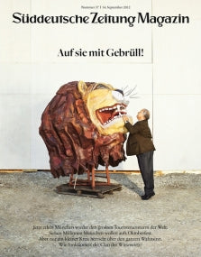 Ein Mann berührt das offene Maul einer großen Löwenskulptur, die an einem sonnigen Tag im Freien steht.