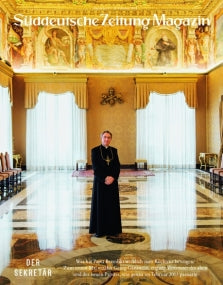 Ein Mann in schwarzer klerikaler Kleidung steht in einem reich verzierten, goldenen Saal mit großen Fenstern und Kronleuchtern.