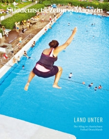 Eine Person in Badekleidung springt in ein großes Freibad, das an einem sonnigen Tag mit Badegästen gefüllt ist.