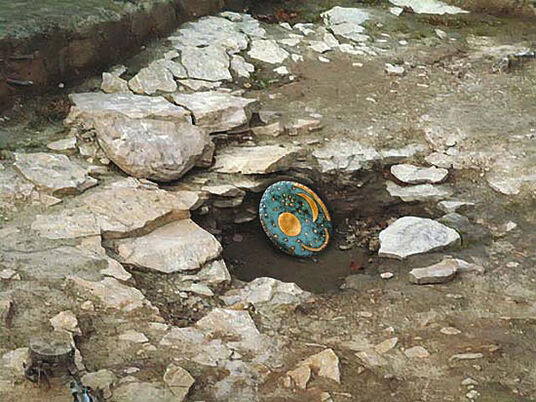 Eine Bronzescheibe mit Goldeinlagen liegt teilweise zwischen Felsen vergraben auf einer archäologischen Ausgrabungsstätte.