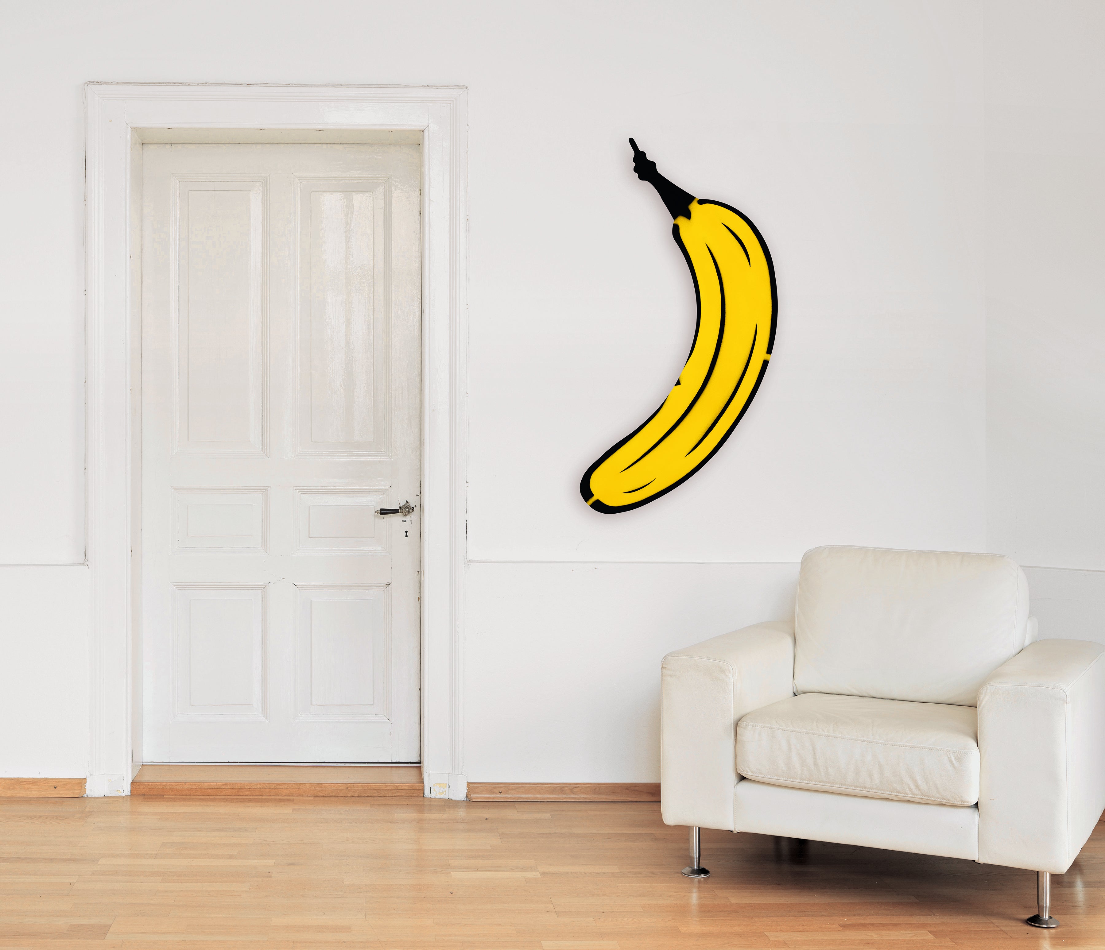 Ein weißer Raum mit einer geschlossenen Tür, einem weißen Sessel und einem großen Bananenkunstwerk an der Wand.