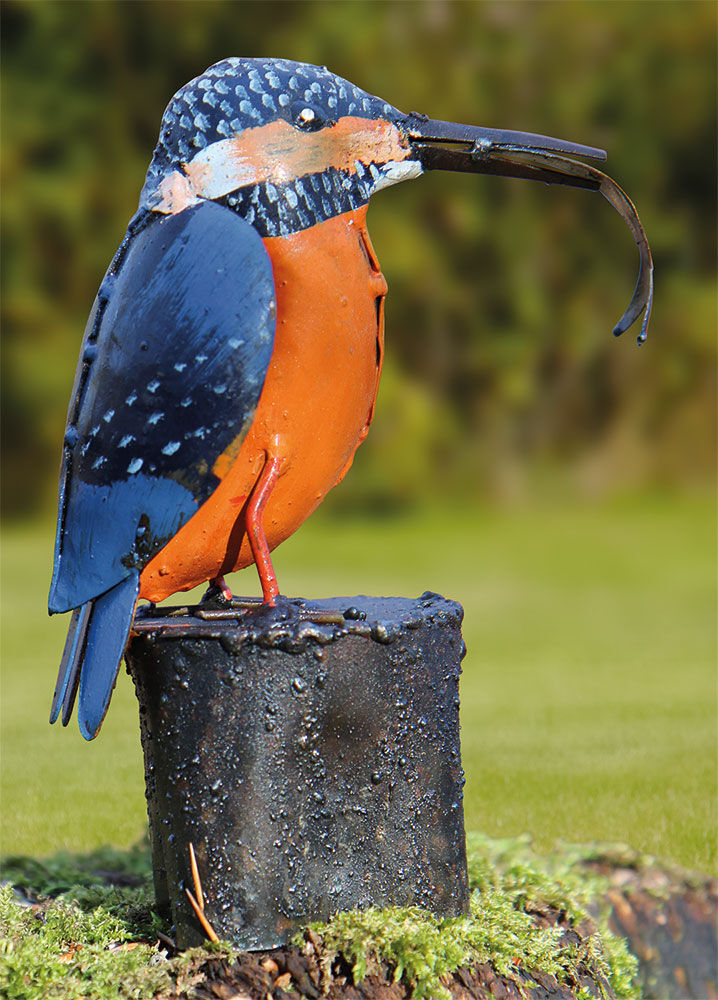 Eine farbenfrohe Vogelskulptur aus Metall steht auf einem schwarzen Sockel im Freien, im Hintergrund grünes Gras.