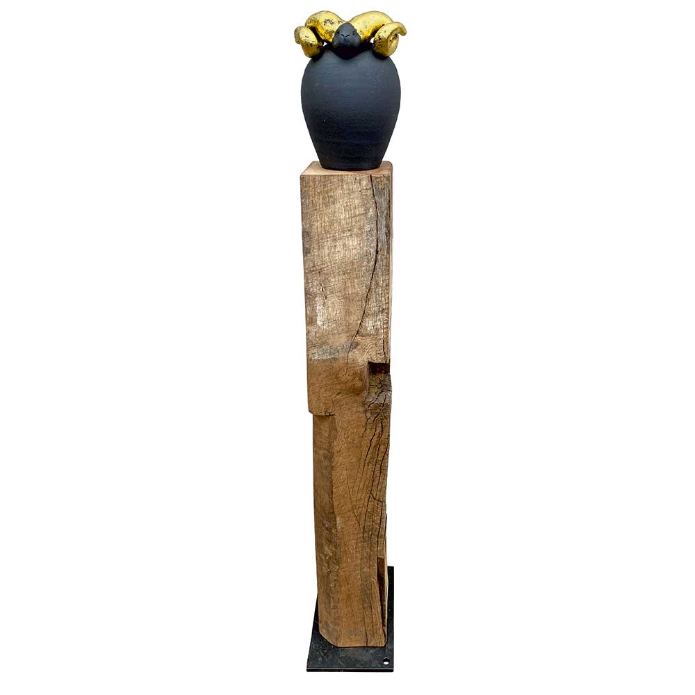 Eine dunkle Vase mit goldenen Bananen auf einem hohen, rustikalen Holzsockel vor weißem Hintergrund.
