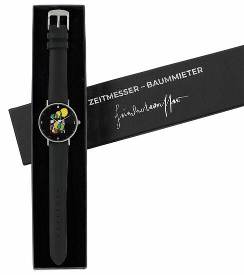 Schwarze Armbanduhr mit abstraktem, buntem Zifferblatt in einer Box mit der Aufschrift ZEITMESSER – BAUMMIETER und einer Signatur.