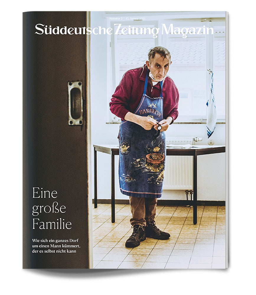 Ein älterer Mann in einer Schürze steht in einer Küche an einem Tisch und schaut mit gefalteten Händen nach unten.