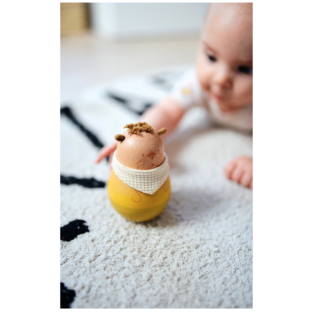 Ein Baby greift nach dem Holzspielzeug Baby Stehaufspielzeug „Seaside“, das auf einem weißen Teppich liegt.