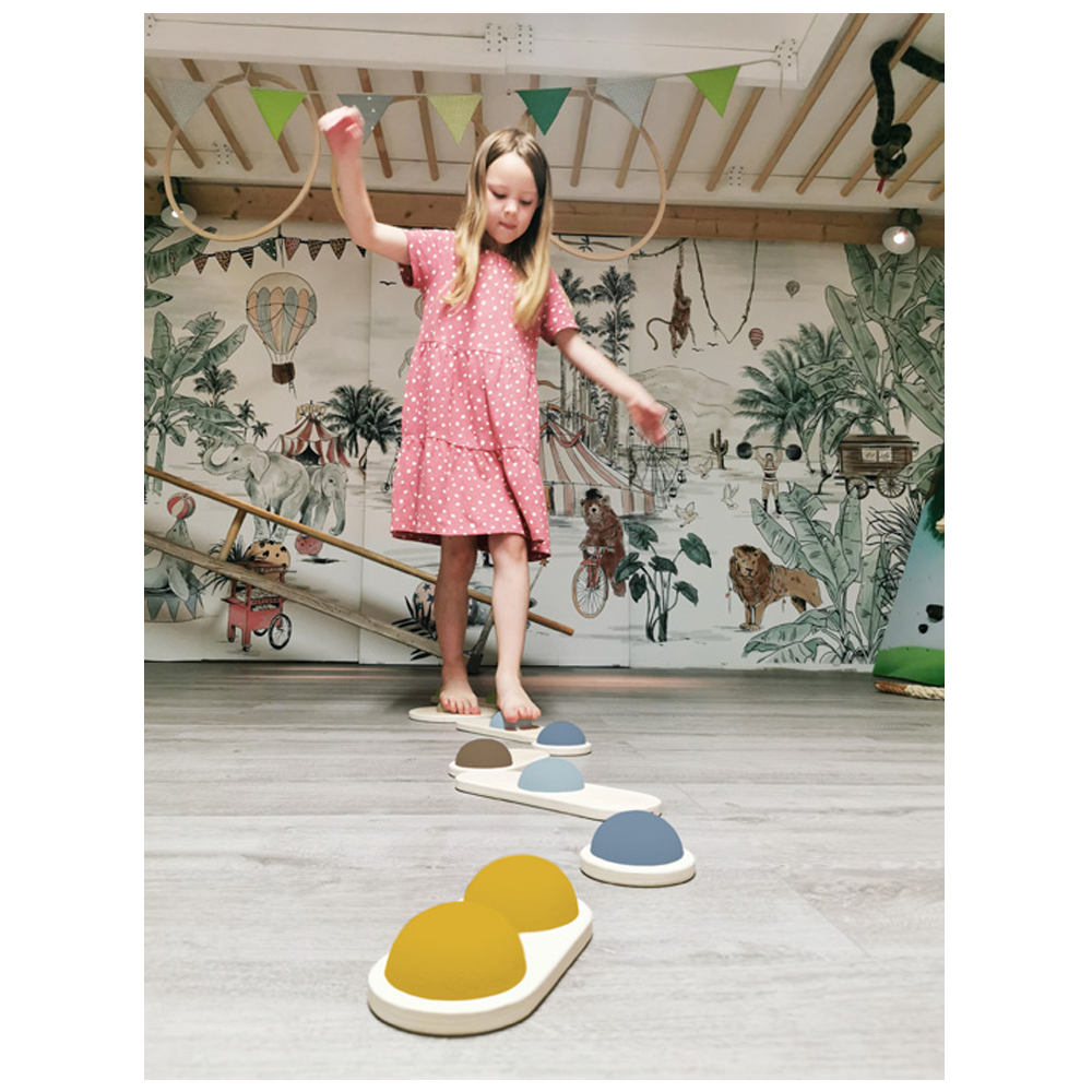 Ein Mädchen in einem rosa Kleid balanciert auf den Balanciersteinen „Adventure“ in einem mit Tier- und Dschungel-Wandkunst dekorierten Spielzimmer.
