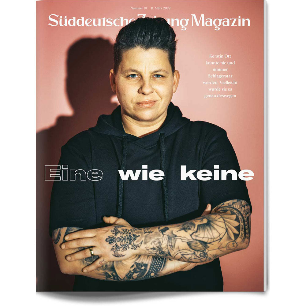 Eine Person mit kurzen Haaren und Tattoos verschränkt die Arme vor einem rosafarbenen Hintergrund auf einem Zeitschriftencover.