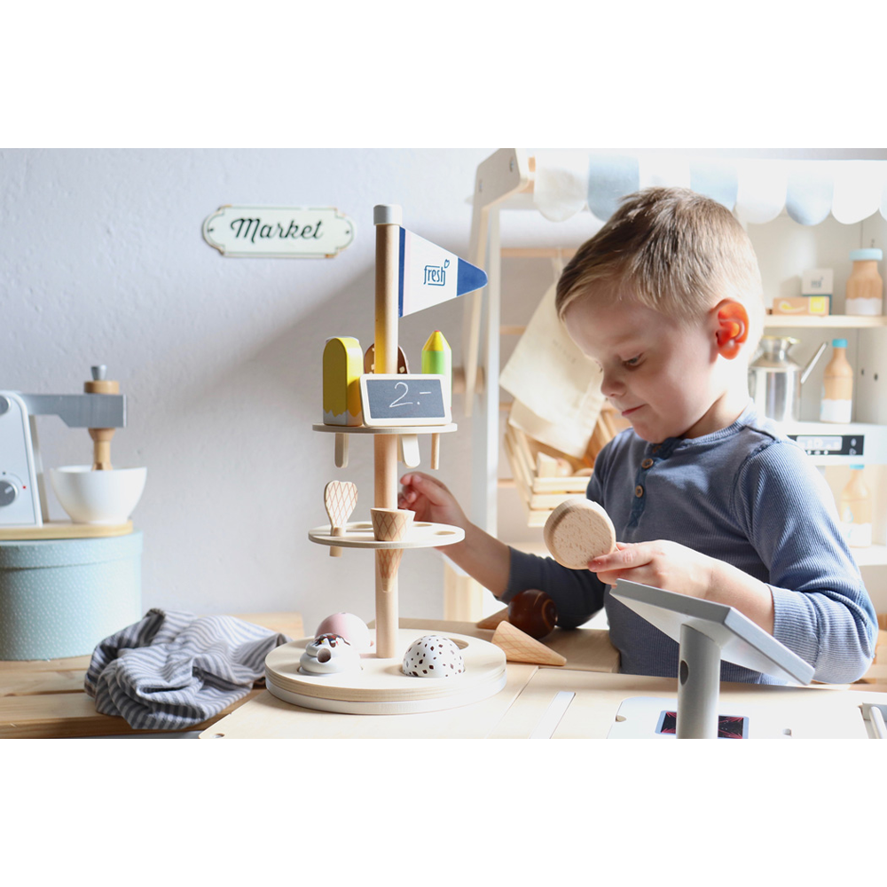 Ein kleiner Junge spielt in seiner Spielküche mit dem Eisständer „Fresh“ aus Holz auf einem Spielmarkt.